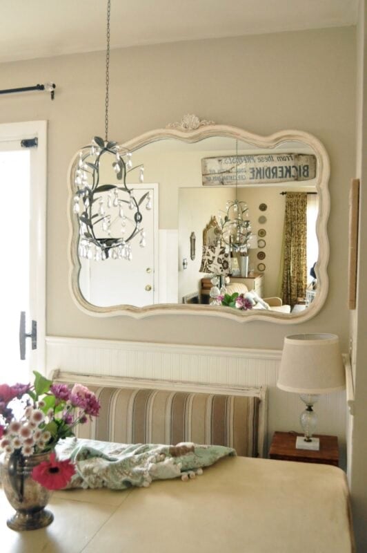 A cozy kitchen nook with a striped bench, floral arrangement, and table lamp sits beneath a large ornate mirror reflecting a chandelier and part of the adjacent room.