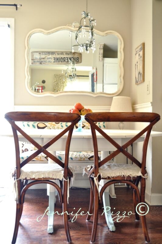 Two wooden chairs with tied seat cushions face a white table. A large mirror above reflects a chandelier and kitchen decor, adding charm to this cozy spot from Our Home Tour.