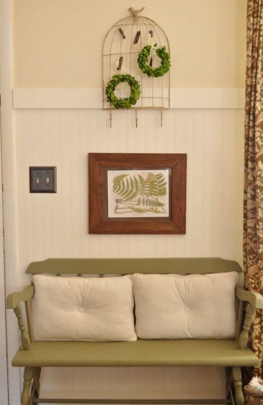 Small green bench with two cream cushions, framed fern artwork, and decorative wall hanging with greenery displayed above on a cream paneled wall.