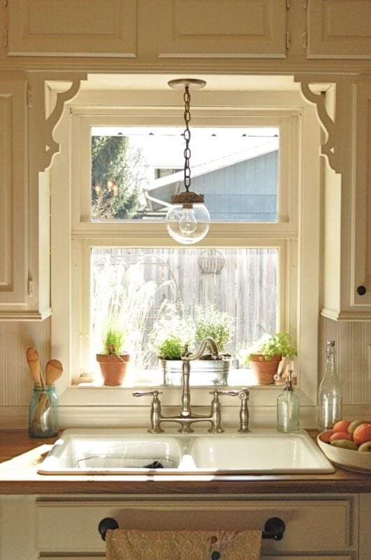 A white farmhouse kitchen sink under a window with potted plants on the sill, a Kitchen Pendant Light Makeover featuring a hanging glass fixture, and wooden utensils on the counter.