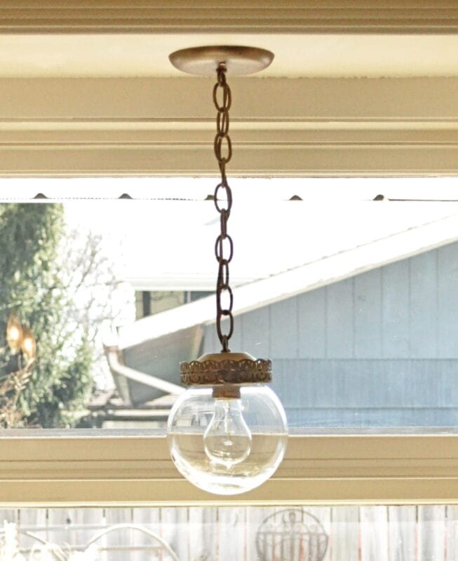 A glass kitchen pendant light fixture with a metal chain hangs from the ceiling in front of a window, showcasing a stunning Kitchen Pendant Light Makeover with an outdoor scene visible in the background.
