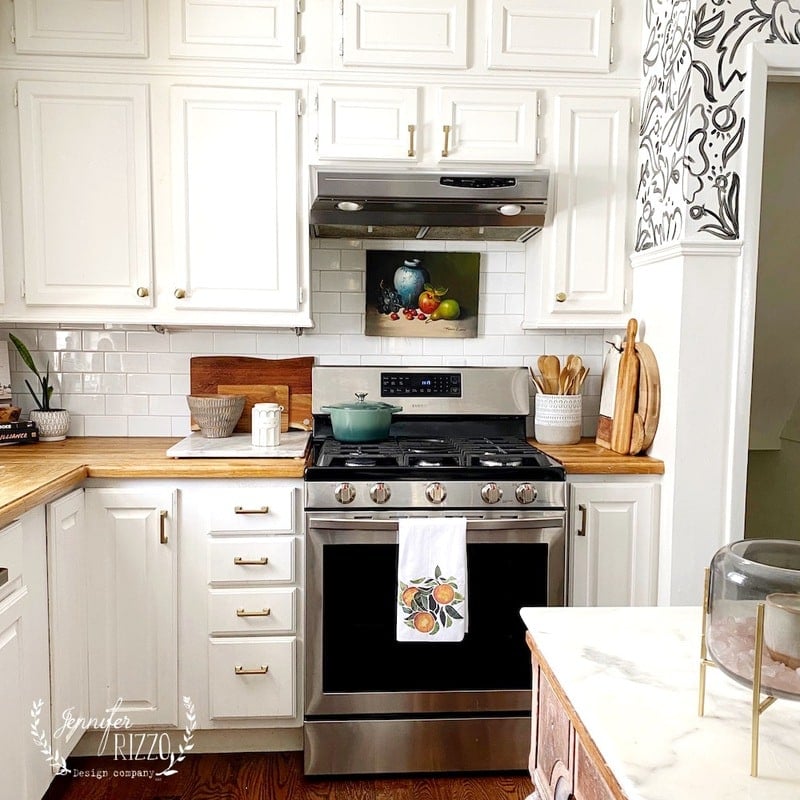 A modern kitchen with white cabinets, stainless steel stove, wooden countertops, and various kitchenware on the counters. A tea towel hangs on the oven door, adding a cozy touch to this space featured in Our Home Tour.
