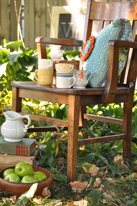A wooden chair with a pillow holds assorted mugs and a small teapot from The Vintage Tea Collection Look Book. Below are stacked books, a white pitcher, and a wooden bowl of green apples, all set outdoors among plants.