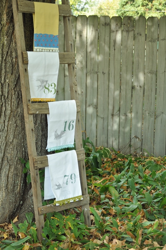 A wooden ladder leans against a tree, showcasing three numbered fabric prints and a yellow towel. The scene, reminiscent of "The Vintage Tea Collection Look Book," features a wooden fence in the background, scattered with autumn leaves.