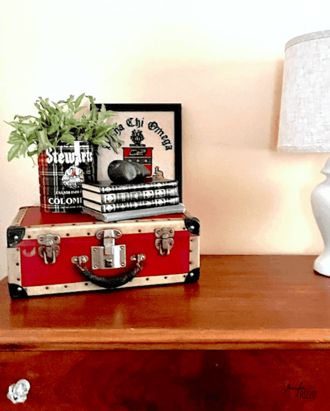 Decorating with vintage suitcases, a red and beige vintage trunk sits on a wooden dresser topped with books, a plaid tin filled with greenery, a framed sign, and part of a white lamp visible on the right.