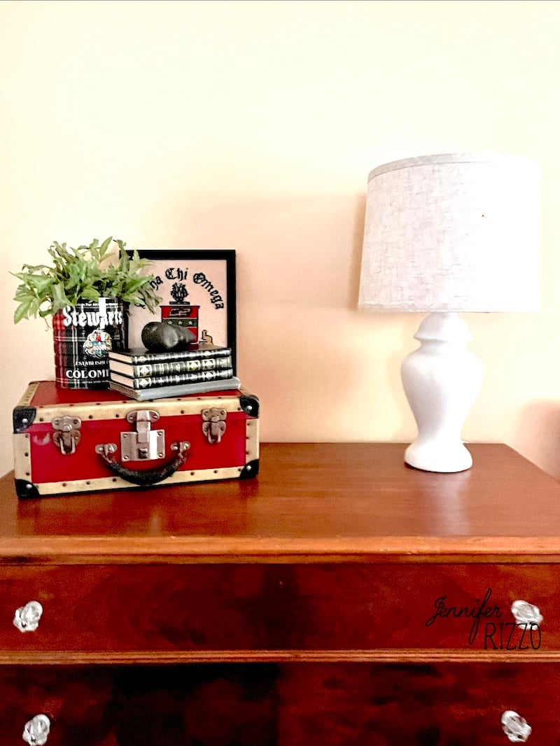 A wooden dresser styled for decorating with vintage suitcases features a white table lamp, a stack of books, a red suitcase, a framed print, and a small plant in a glass bottle.