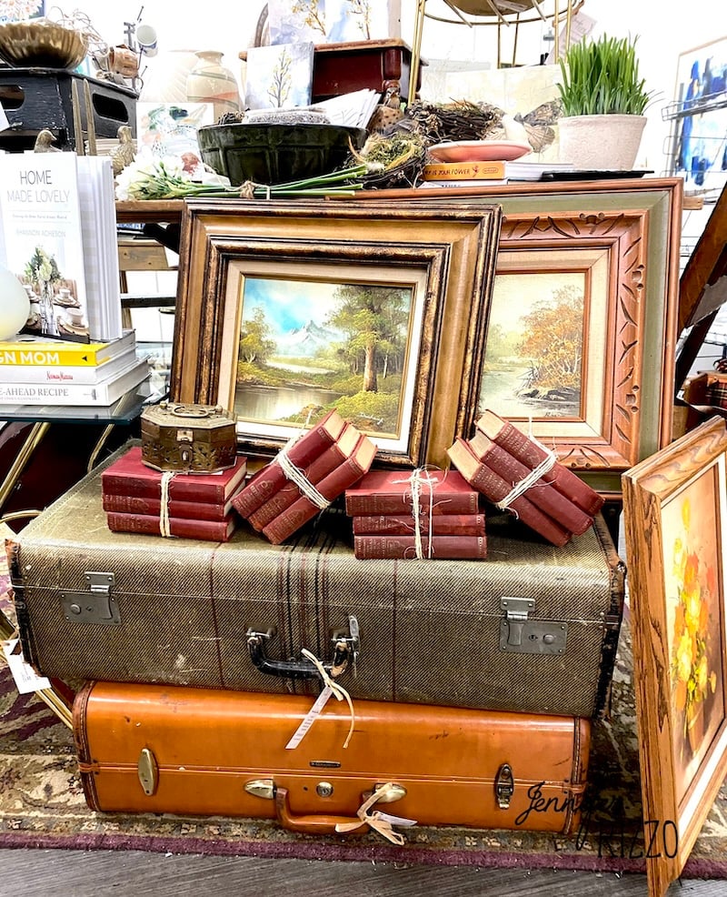 A stack of vintage suitcases topped with small, tied red books and framed landscape paintings creates a charming display—perfect inspiration for decorating with vintage suitcases among your favorite home decor items.