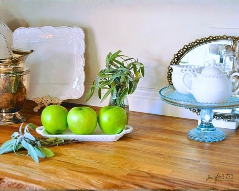 Three green apples in a white dish, fresh sage, a silver pitcher, square plates, and a teapot on a glass stand are arranged on wood countertops after 4 years—still looking charming and timeless atop the wooden surface.