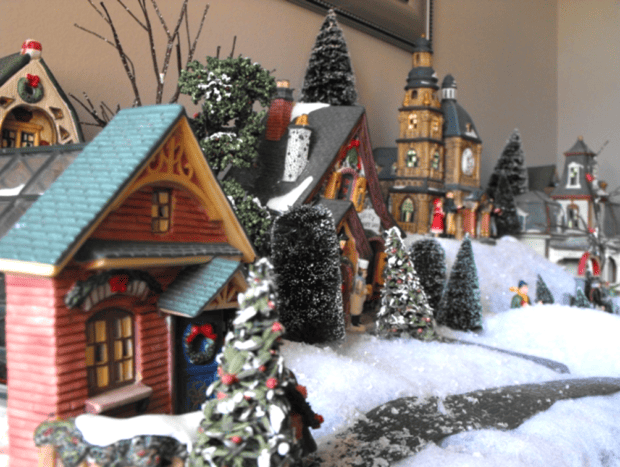 Miniature winter village scene with snow-covered model houses, trees, and small figurines arranged on a tabletop, capturing the charm of vintage and cozy Christmas decor.