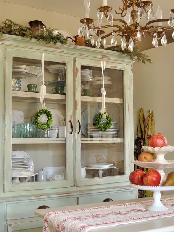 A vintage glass-front cabinet displays neatly arranged dishes and glassware, with small wreaths on the doors. Touches of red in a holiday kitchen tour appear with a fruit stand filled with apples sitting on the table in the foreground.