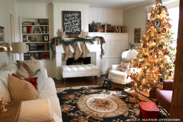 A living room decorated with vintage and cozy Christmas decor features a lit tree, stockings on a fireplace mantel, a white sofa, armchair, bookshelves, and a floral rug.