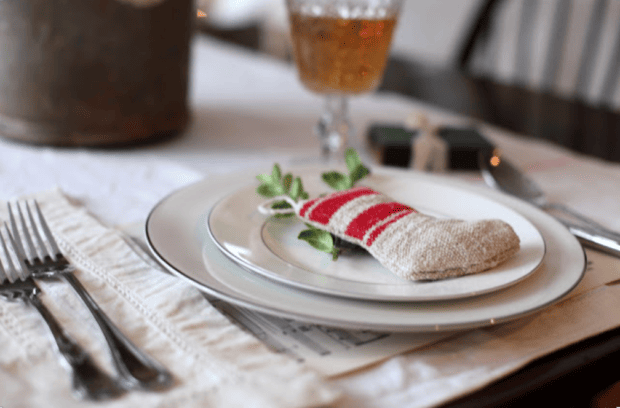 A table setting with a white plate, silverware, and a small red-and-white striped stocking decoration on the plate captures the spirit of Holiday Housewalk 2011 Day 2 with its charming traditional holiday decor. A gift and glass of beverage are in the background.