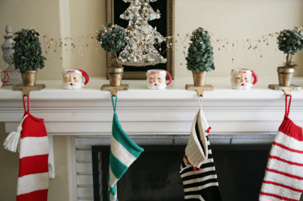 Four Christmas stockings hang from a white mantel, each held by a Santa head stocking holder. Small potted faux trees and a beaded tree decoration create vintage and cozy Christmas decor atop the mantel.
