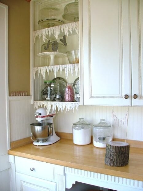 White kitchen with glass-front cabinet displaying dishes, a mixer on the wooden counter, jars with flour and sugar, and a wooden log decoration—perfect for a cozy holiday housewalk.
