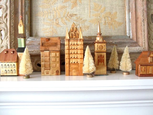Wooden miniature buildings and small bottle brush trees are arranged in a row on a white mantel, creating a charming holiday housewalk scene with textured framed artwork in the background.