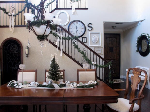 A dining room decorated for Christmas, perfect for a holiday housewalk, with a garland on the table, a small tree centerpiece, garlands on the stair railing, and white ribbons throughout.