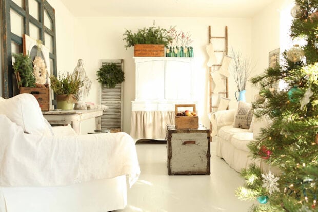 Bright living room decorated for Christmas with a white armchair and sofa, vintage furniture, a Christmas tree, and festive greenery—perfect for a holiday housewalk filled with ornaments and seasonal cheer.