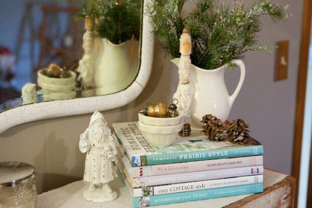 A stack of home decor books topped with a Santa figurine, pinecones, a bowl of ornaments, and a white pitcher with greenery creates a festive Holiday Housewalk vibe near the mirror on the table.