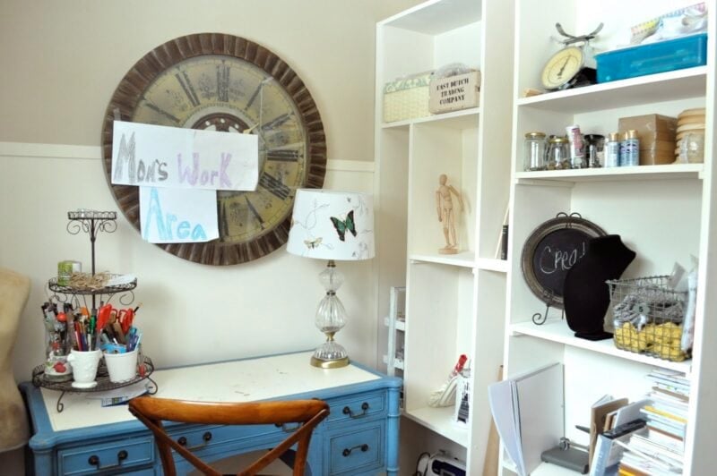 A home office setup with a blue desk, art supplies, a lamp, a large clock, shelves with craft items, a built-in bookcase, and a sign reading "Mom's Work Area.