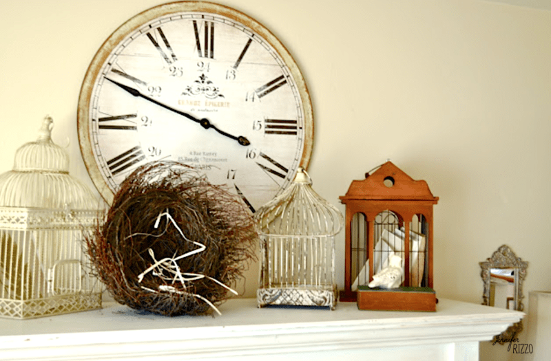 A large vintage-style wall clock is displayed above a mantel decorated with birdcages, a bird’s nest, and a small brown house-shaped ornament, creating a refreshed after holiday living room vibe.