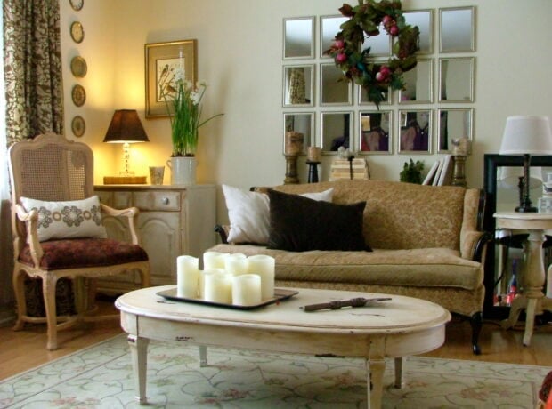 A living room with vintage-style furniture, a beige sofa, an armchair, a coffee table with candles, wall decor, and a mirror with a wreath above the sofa.
