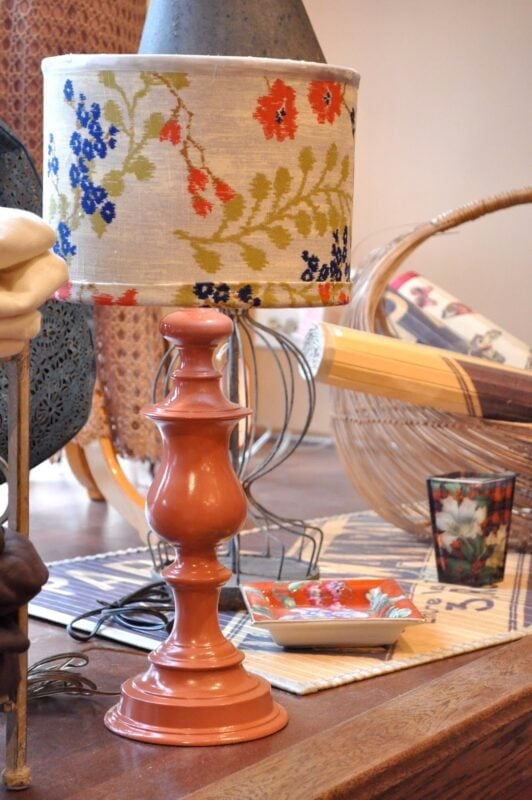 A ceramic table lamp with a floral-patterned lampshade, fresh from a lamp makeover with spray paint, sits on a wooden surface surrounded by decorative items including a glass, a dish, and a fan.