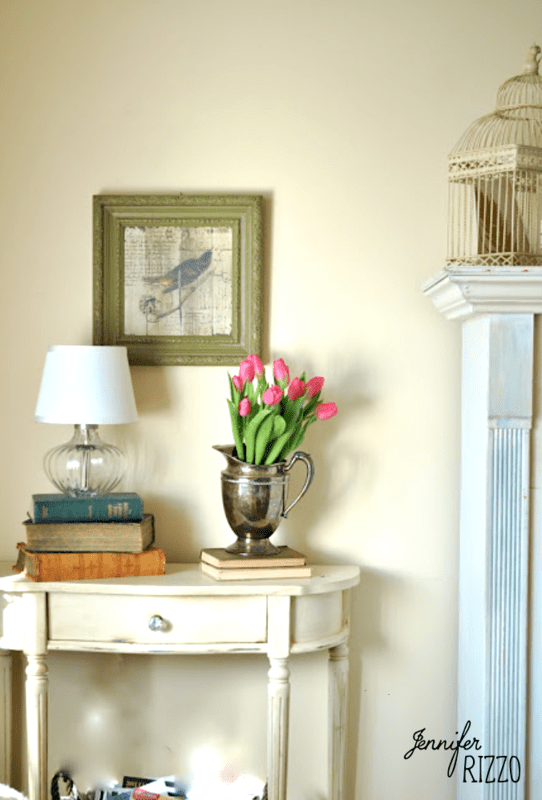 A small table with stacked books, a lamp, and a silver vase of pink tulips adds charm to this refreshed after holiday living room; framed bird art and a decorative birdcage adorn the wall above.