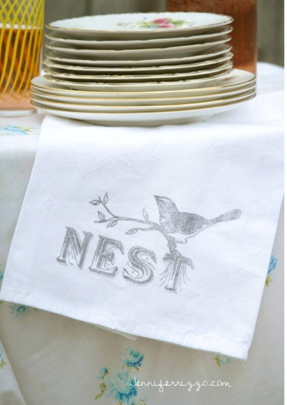 A white cloth from the Sparrow and Blossom Collection, with a bird and the word "NEST" printed on it, hangs over a table edge in front of a stack of floral plates and a yellow basket.