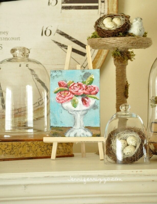 A small painting from the Sparrow and Blossom Collection, featuring pink flowers on an easel, is displayed on a shelf with glass domes covering bird nests and eggs, next to a clock face in the background.