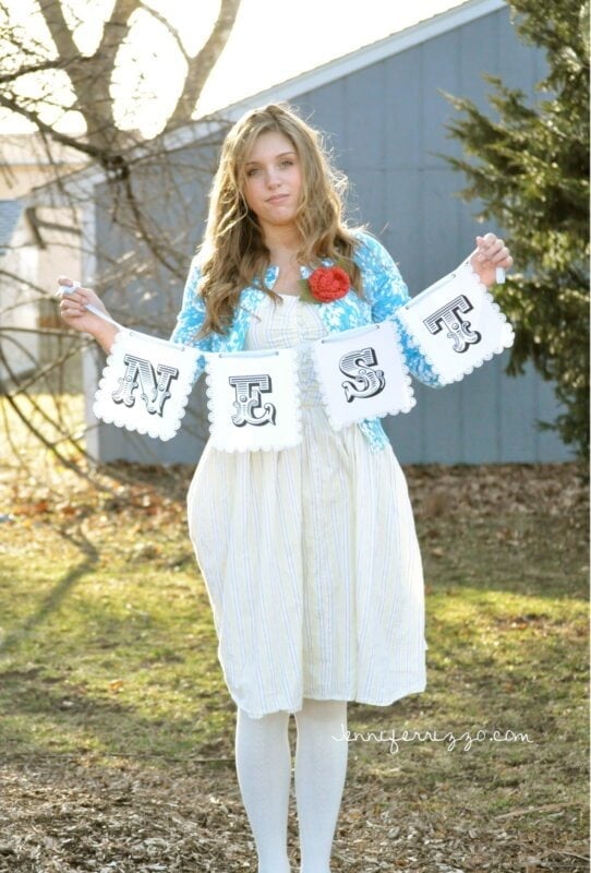 A woman stands outdoors holding a banner that spells "NEST" with large black letters, in front of a blue building and trees, celebrating the Sparrow and Blossom Collection.