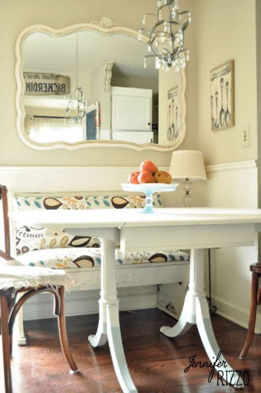 A small dining nook with a white table, patterned cushions, a bowl of fruit, a chandelier, and a large ornate mirror on the wall highlights subtle accents using the Dip Dye Paint Technique for added charm.