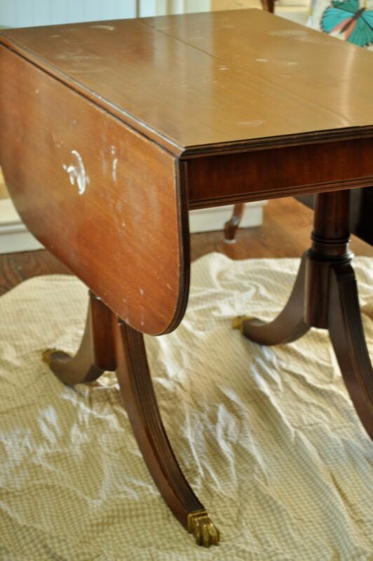 A wooden drop-leaf table with two curved legs on each side, featuring a subtle dip dye paint technique, is placed on a textured cloth on a hardwood floor.