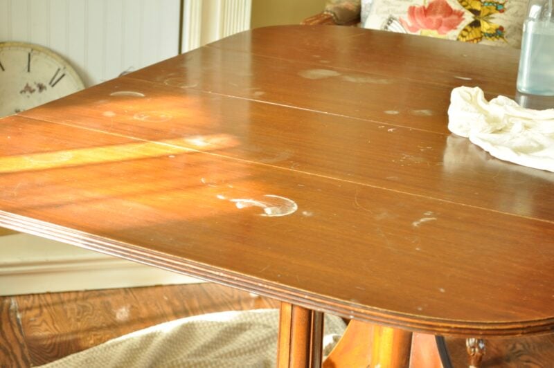 A wooden table with noticeable white water stains and a cloth placed on one corner, illuminated by sunlight, awaits a refresh with the Dip Dye Paint Technique.