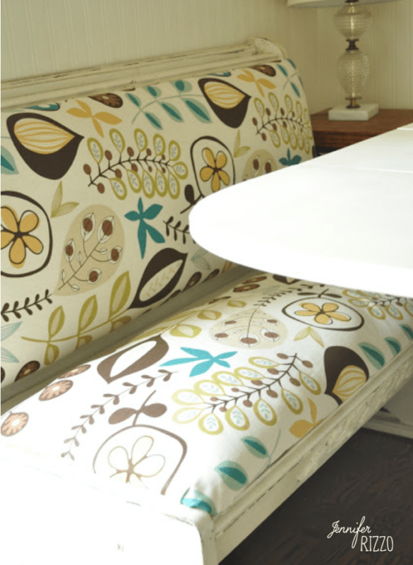 A bench with retro floral patterned upholstery in shades of yellow, green, and brown sits beside a white oval table; a lamp is visible in the background, while dip dye paint technique accents add a modern touch to the decor.