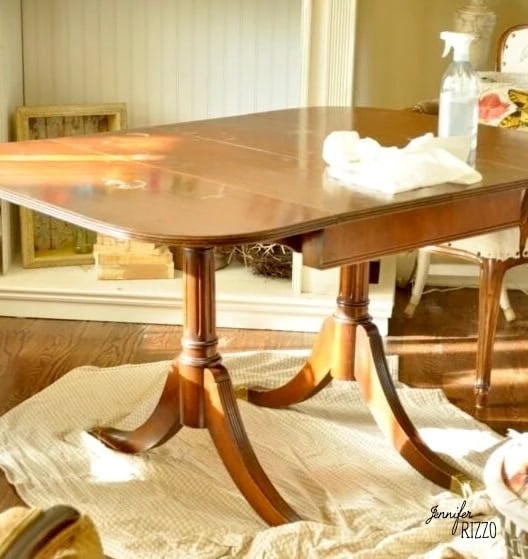 A wooden dining table is set on a rug with a cleaning cloth and spray bottle placed on top, suggesting the table is being cleaned, perhaps after trying out the Dip Dye Paint Technique for a fresh new look.