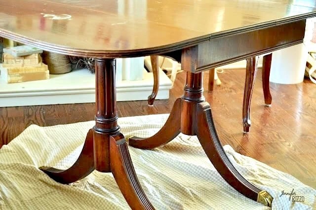 A dark wooden dining table with double pedestal legs stands on a checkered cloth, its base enhanced by a subtle Dip Dye Paint Technique, with books and decor visible in the background.