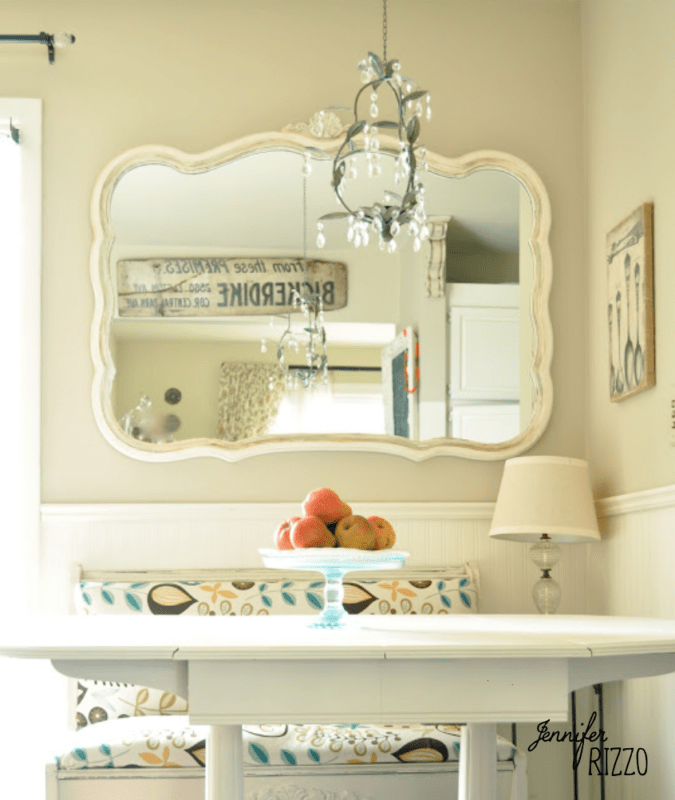A kitchen nook with a white table, a fruit bowl, a cushioned bench, wall art using the dip dye paint technique, a table lamp, and a large ornate mirror reflecting the opposite wall and window.