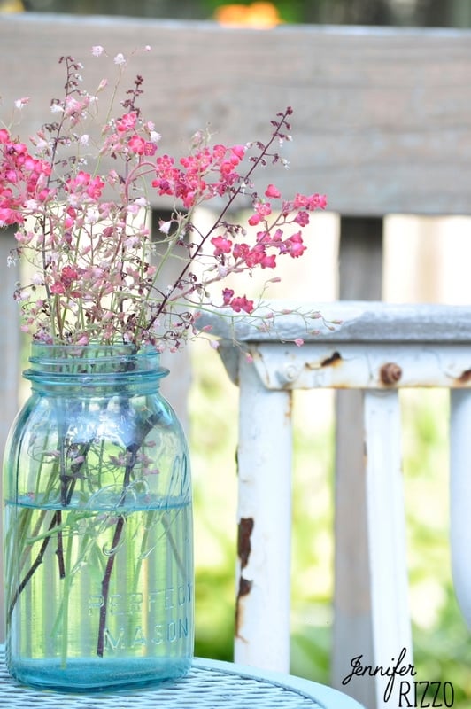 A blue mason jar with pink flowers sits on a weathered white metal chair, perfect for a spray paint outdoor furniture makeover, with a wooden bench in the background.