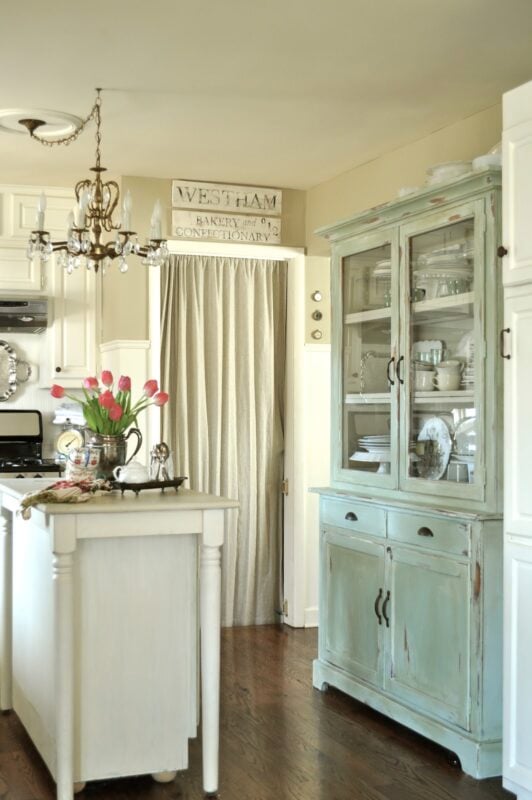 A vintage-style kitchen with a small island, a chandelier, a distressed light blue china cabinet, and a vase of pink tulips. White cabinetry and dishware are visible.