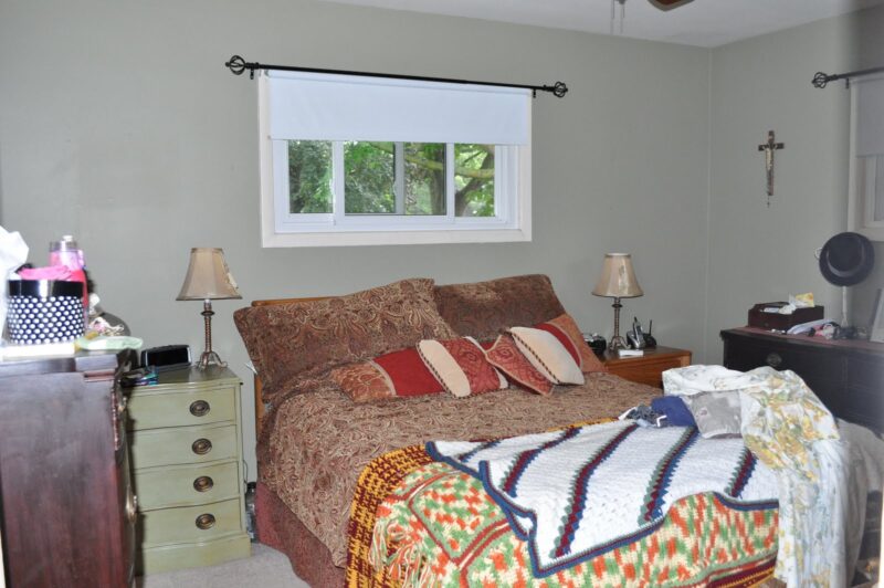 A neatly made bed with patterned pillows and a blanket sits against a wall under a window; nightstands, lamps, and a dresser with various items are in the room.