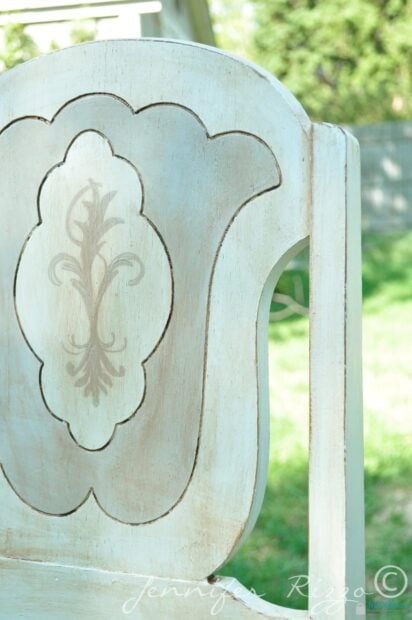 A close-up of a vintage gray and white painted chair with a decorative, painted floral design on the backrest, set outdoors with grass and trees in the background.