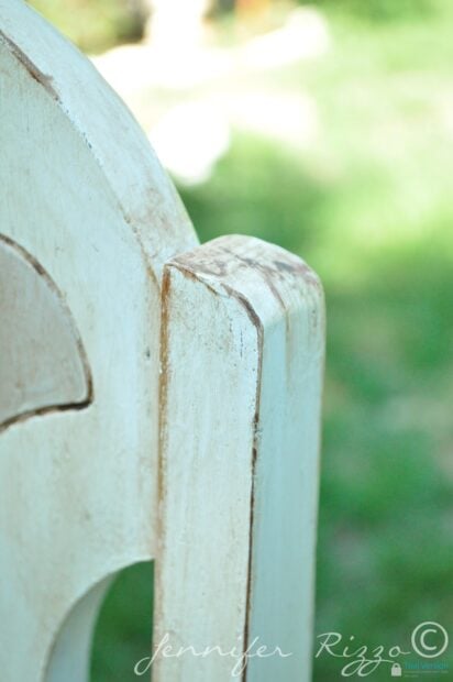 Close-up of the top corner of a distressed, gray and white painted chair with visible wear, set against a blurred green outdoor background.