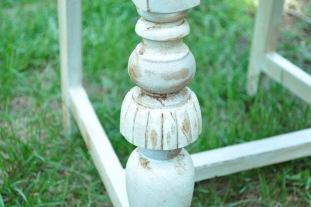 Close-up of a distressed, white-painted wooden table leg with carved details, set on green grass beside a gray and white painted chair.