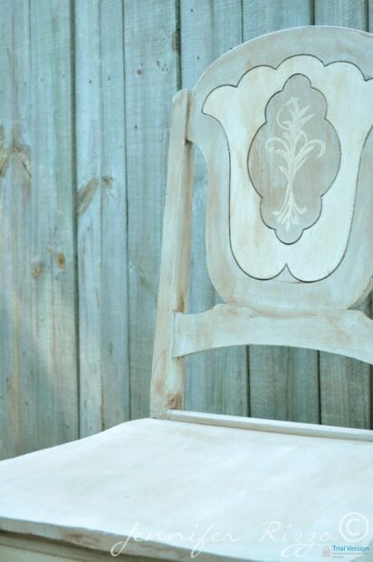A close-up of a gray and white painted chair with carved decorative details on the backrest, positioned in front of a wooden fence.
