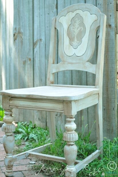 A vintage gray and white painted chair with decorative carvings and distressed details sits on a brick patio near a wooden fence.