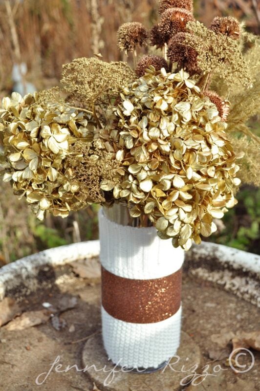 A vase with a white and brown textured design holds a spray painted floral arrangement of dried hydrangeas and other dried flowers, placed on an outdoor surface.
