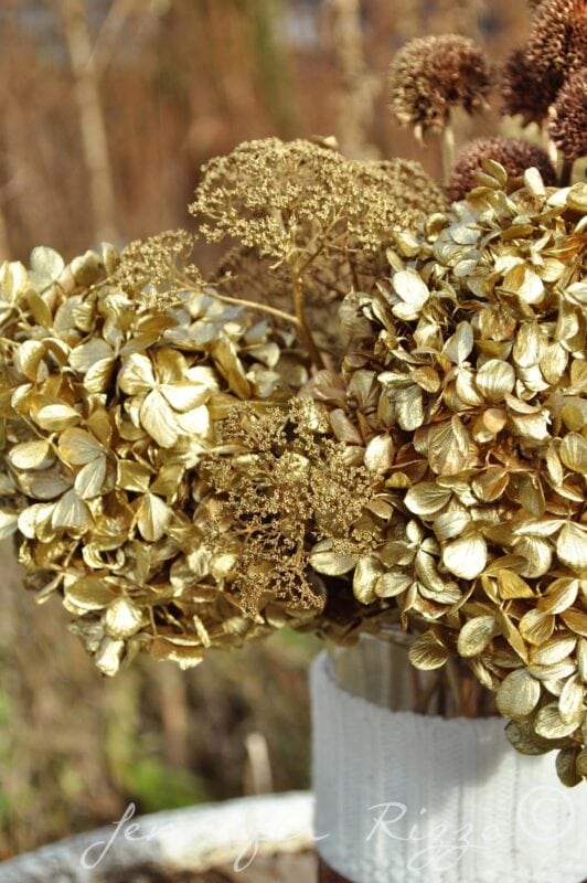 A spray painted floral arrangement of dried hydrangeas and other flowers in gold hues, artfully displayed in a white textured vase.