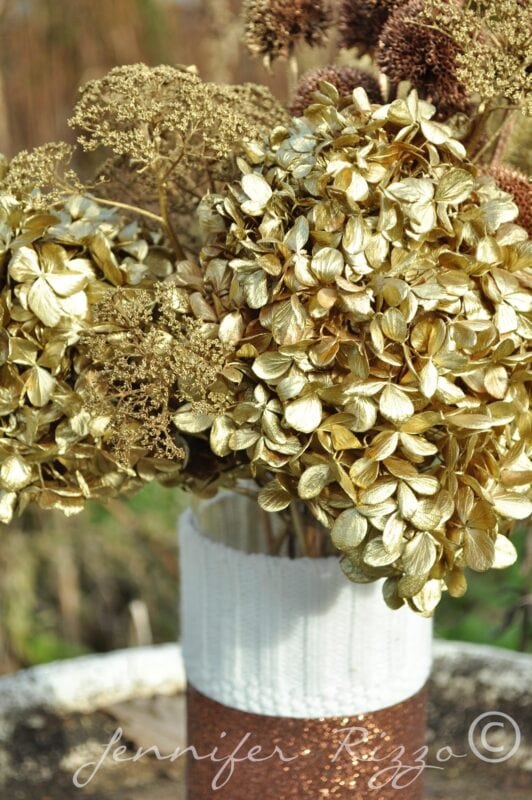 A vase holds a spray painted floral arrangement of dried flowers, including hydrangeas painted gold. The vase is wrapped with white and brown textured materials.