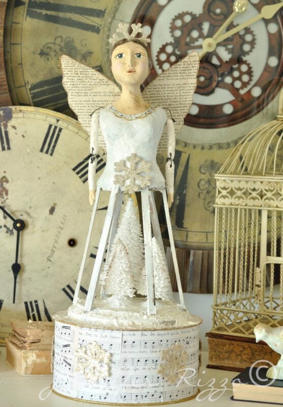 A mixed media Angel Santos Cage Dolls figurine with paper wings and a wire skirt adorned with sheet music, standing before vintage clock faces and a birdcage—perfectly paired with handmade earrings for a unique display.