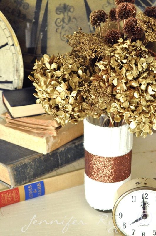 A spray painted floral arrangement in a vase sits on a table near stacked books and two vintage-style clocks.